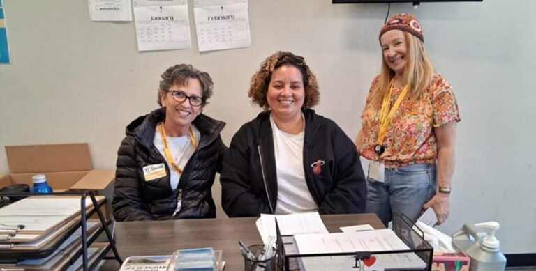 Three women smiling, two seated at a desk with papers, one standing, all wearing name tags in an office setting.