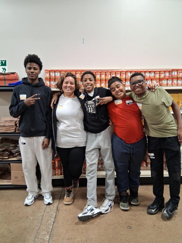 Five people stand smiling together in front of shelves of canned goods, arms around each other in a grocery store.