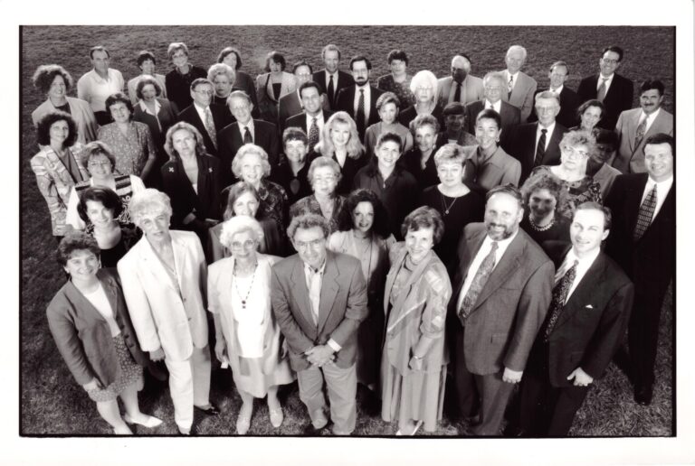 A group of men and women in formal attire stand closely together, smiling and looking up at the camera outdoors.
