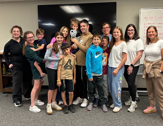 A group of people, including children and adults, smile together indoors in a casual classroom setting.