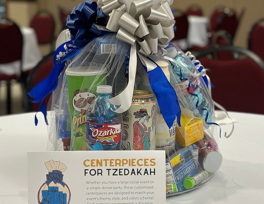 A gift basket with snacks and drinks, wrapped in tulle and bows, sits behind a sign reading "Centerpieces for Tzedakah.