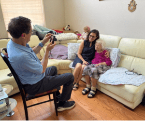 A man takes a photo of two women sitting together on a cream couch in a cozy, well-lit living room.
