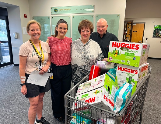 Four smiling adults stand beside a cart filled with Huggies diapers and other donated baby supplies inside a building.
