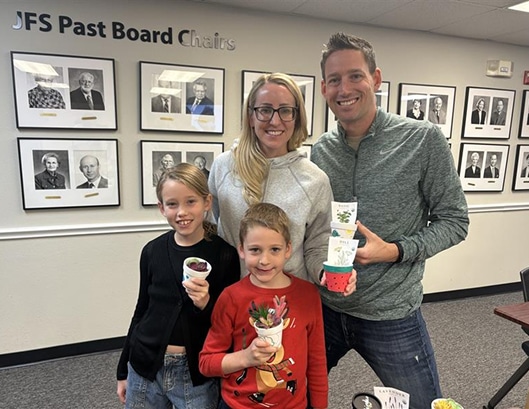 A smiling family of four poses indoors, each holding decorated paper cups, with framed photos on the wall behind them.