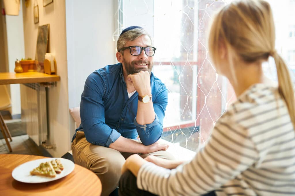 Man in glasses smiling and talking with a woman at a cozy café, with food on the table in front of them.