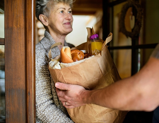 An older woman smiles as someone hands her a paper bag full of groceries at her door.