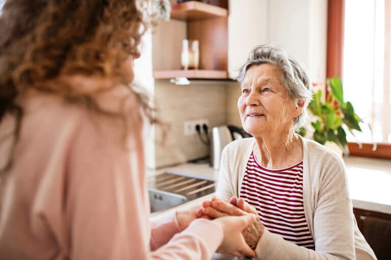 An elderly woman smiles warmly, holding hands with a younger person in a cozy, sunlit kitchen.