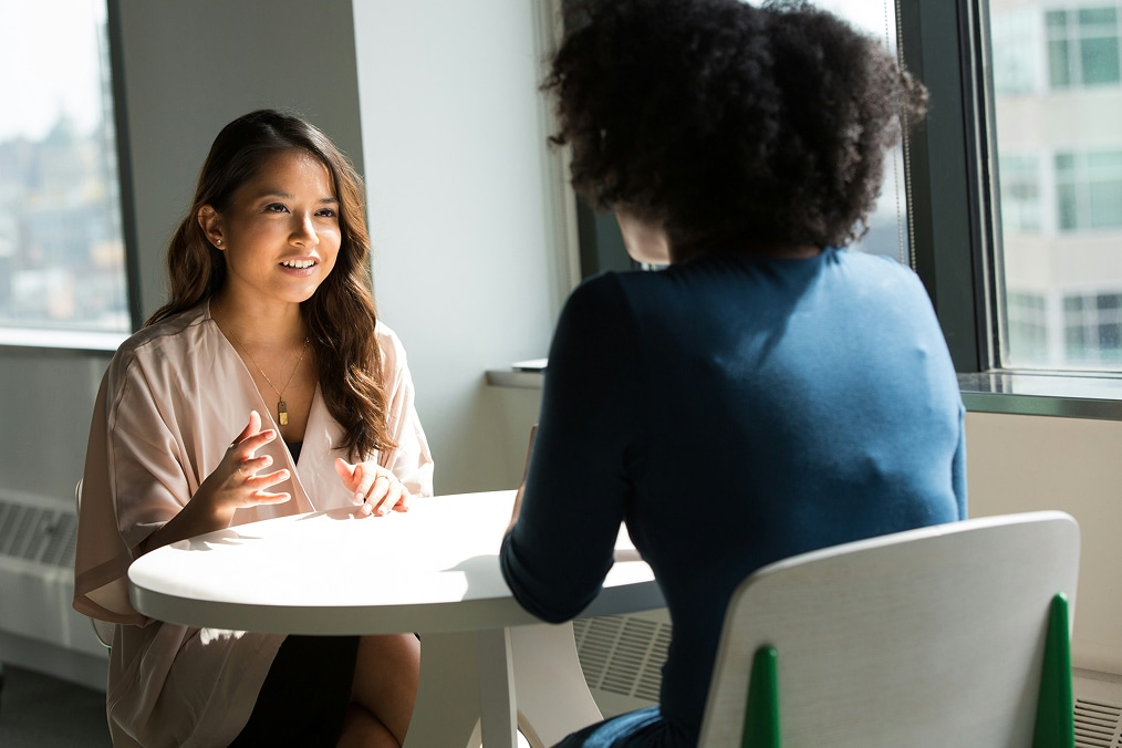 Two women sit across from each other at a round table, having a conversation in a bright office setting.