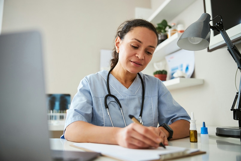 Female doctor in scrubs writes on a clipboard at her desk, with a stethoscope around her neck.