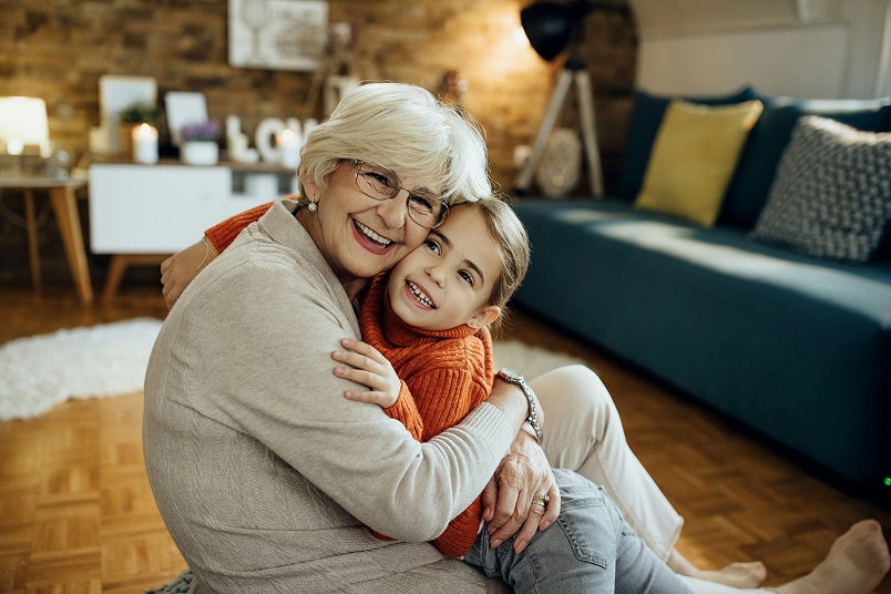 Smiling elderly woman and young girl hugging while sitting on a living room floor, with cozy decor in the background.
