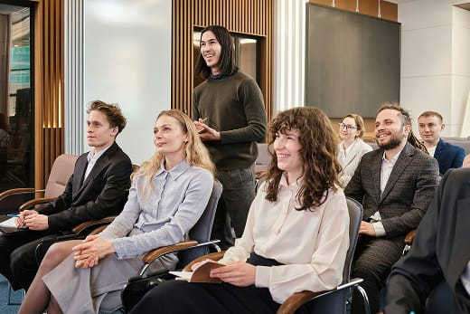 A group of people in business attire sit and smile, while one person stands and speaks in a modern conference room.