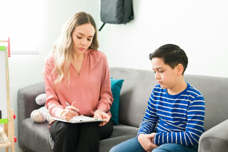 A woman takes notes on a clipboard while a boy in a striped shirt sits beside her, looking down.