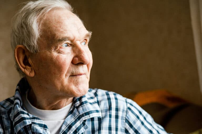 Elderly man with white hair in a plaid shirt looks thoughtfully out of a window with soft natural light on his face.
