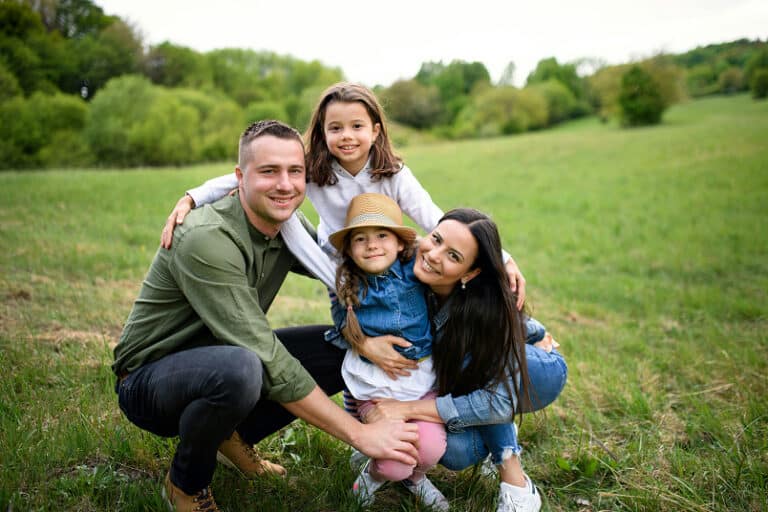 A smiling family of four poses together in a green field, with trees and grass in the background.