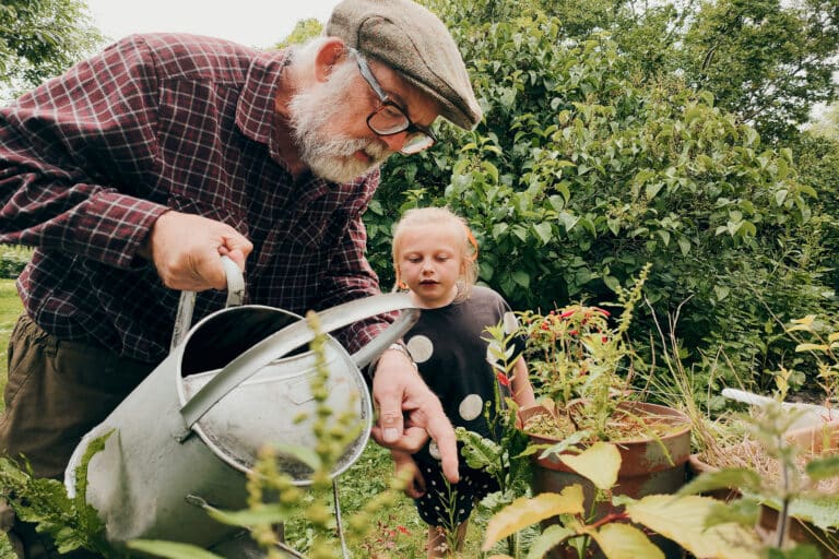 An elderly man and a young girl water plants together in a lush, green garden.