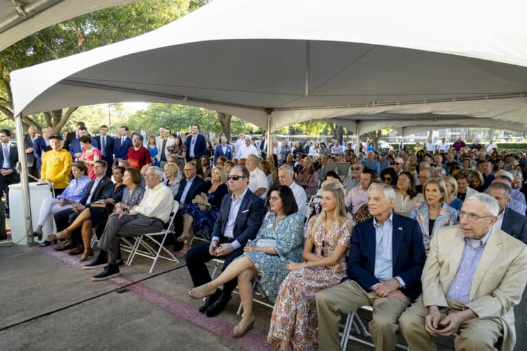 A large group of people sits under white tents at an outdoor event, attentively watching something ahead.