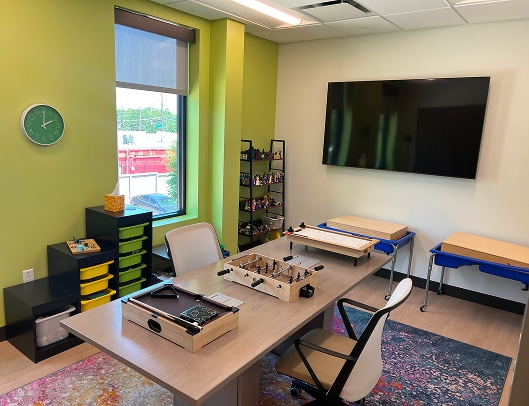 Bright office with green walls, large TV, desk, games, storage bins, and two light blue therapy tables near the window.
