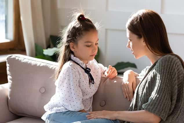 Woman comforts a young girl on a couch, both looking serious and engaged in conversation.