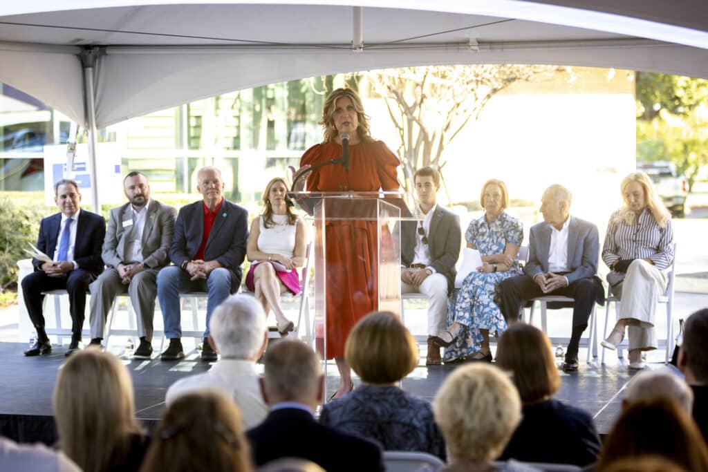 A woman speaks at a podium on stage, with a seated panel and audience at an outdoor event under a tent.