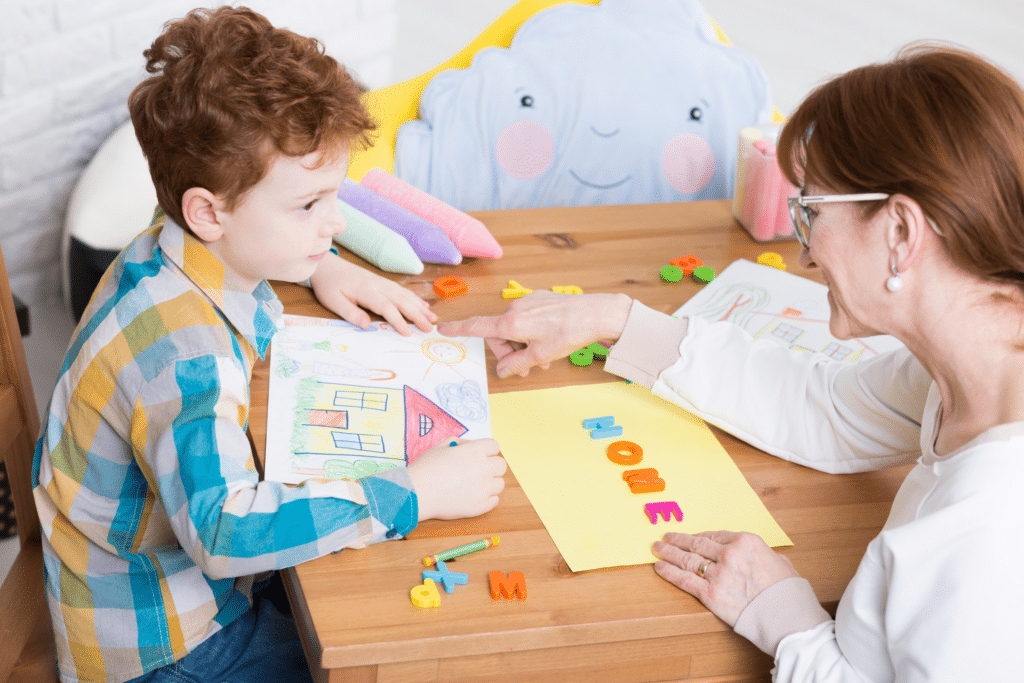 A woman and a young boy sit at a table, drawing and spelling “home” with letters, surrounded by colorful art supplies.
