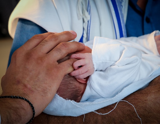 A newborn baby wrapped in a white blanket is lovingly touched on the head by an adult's gentle hand.