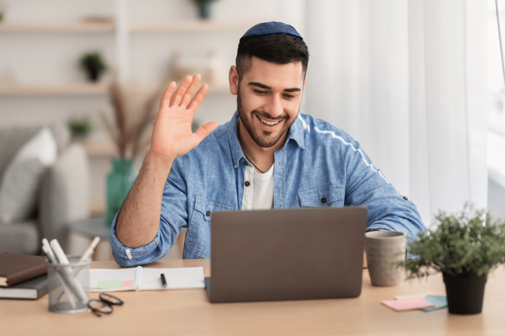 Smiling man in a blue shirt waves at his laptop during a video call, sitting at a desk with notebooks and a mug.