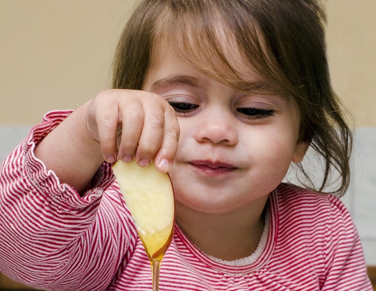 Young child in a red striped shirt dipping an apple slice into honey, smiling and looking down at the food.