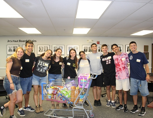 A group of smiling young people stands behind a shopping cart full of groceries and toys in an indoor room.