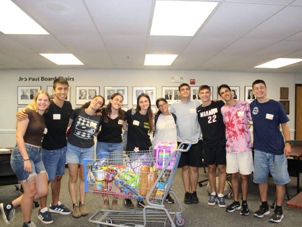 Ten smiling young adults stand together behind a shopping cart filled with groceries in a room with framed photos on the wall.