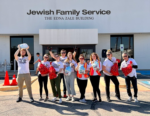 Eight smiling people stand in front of Jewish Family Service holding bags and food items, ready to help distribute meals.