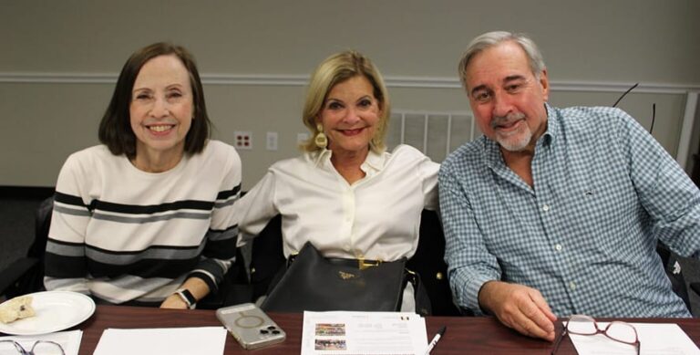 Three smiling adults sit at a table with papers, a phone, and glasses in front of them, posing for the camera indoors.
