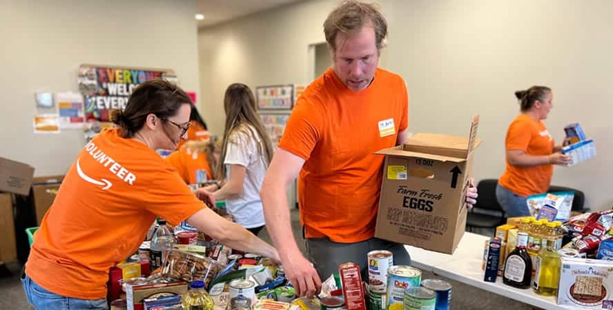 Volunteers in orange shirts sort and pack food items at a donation center with tables full of canned goods and groceries.
