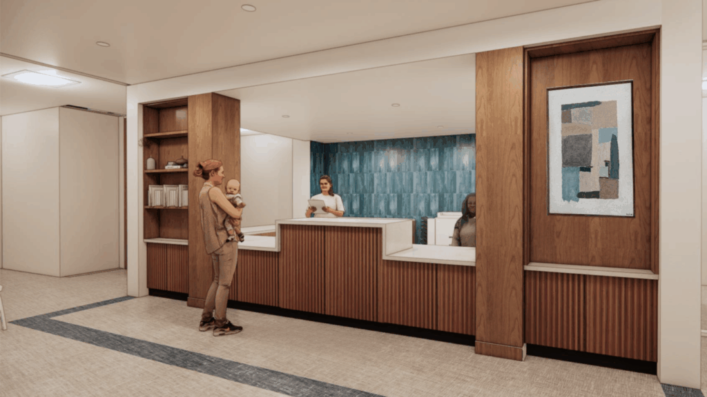 A woman holding a baby stands at a modern reception desk with two receptionists and abstract art on the wall.