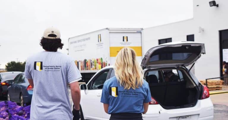Two volunteers in matching shirts stand by a car with its trunk open at a food distribution event.