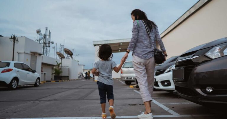 A woman and a young girl holding hands walk through a parking lot lined with cars on a cloudy day.