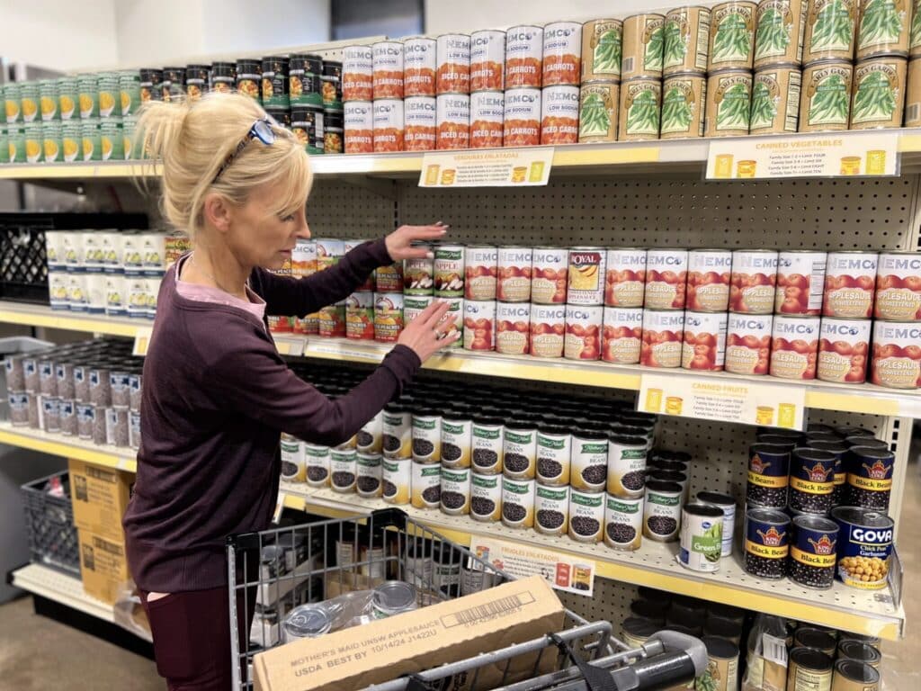 Woman in a grocery store aisle selecting canned goods from a shelf, with a shopping cart in front of her.