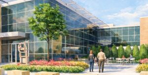 Two people walk toward a modern glass building surrounded by flowers, trees, and outdoor seating on a sunny day.