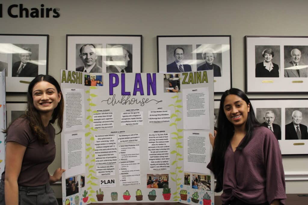 Two young women stand smiling in front of a colorful display board labeled “PLAN Clubhouse,” with portraits on the wall behind them.