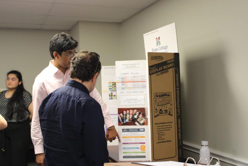 Two people stand by a display board, discussing its content; another person stands in the background in a classroom.