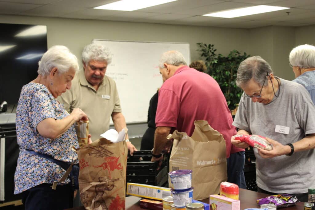Seniors sort food items into bags around a table in a community room with a whiteboard and plants in the background.