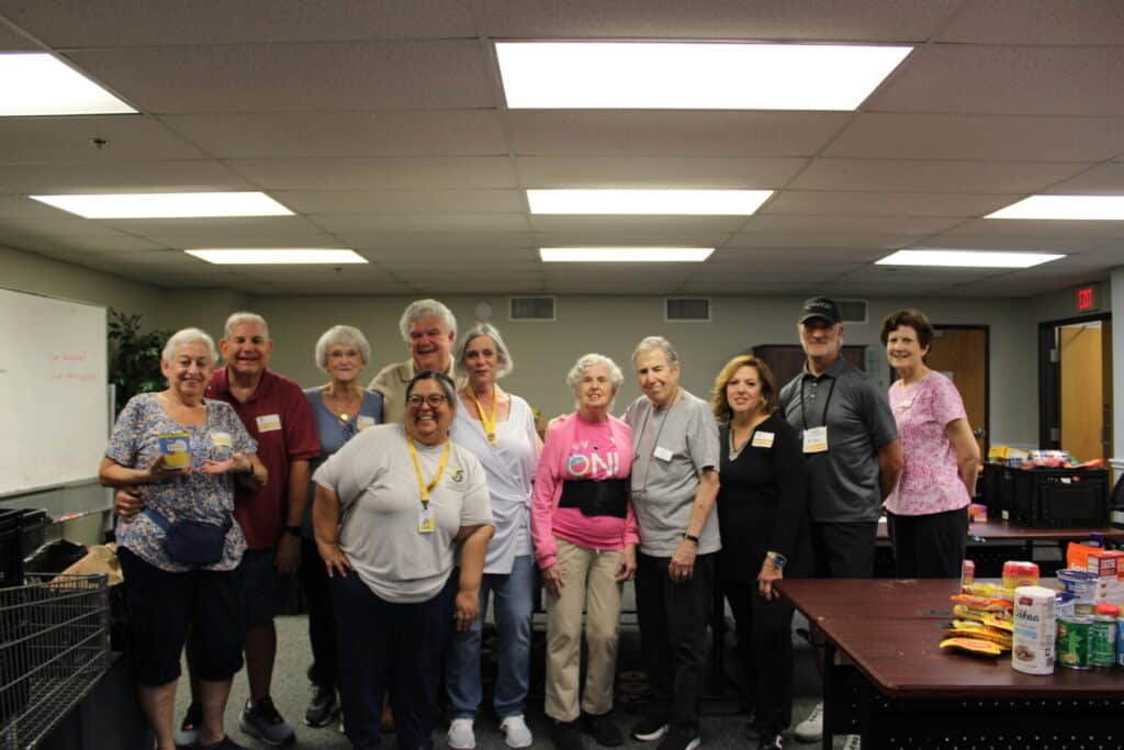 A group of smiling adults stands together in a room with food items and supplies on tables.