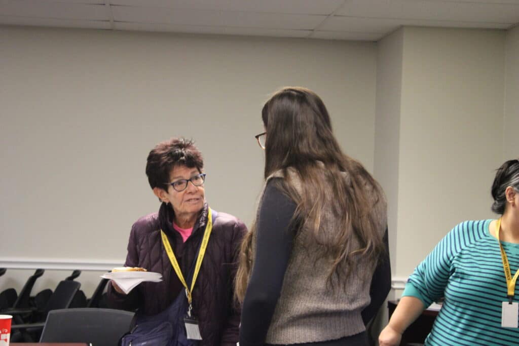 Two women with lanyards talk in an office; one holds a plate, while another stands with her back to the camera.