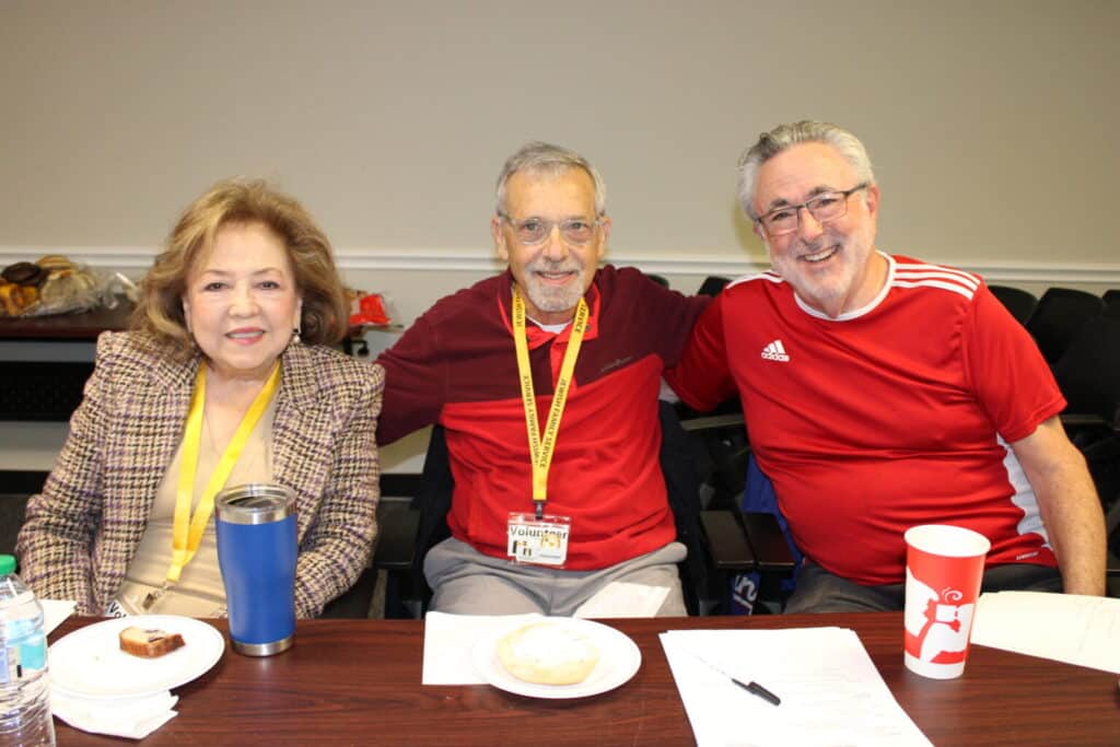 Three smiling adults sit together at a table with drinks and snacks, papers in front of them, posing for a group photo.