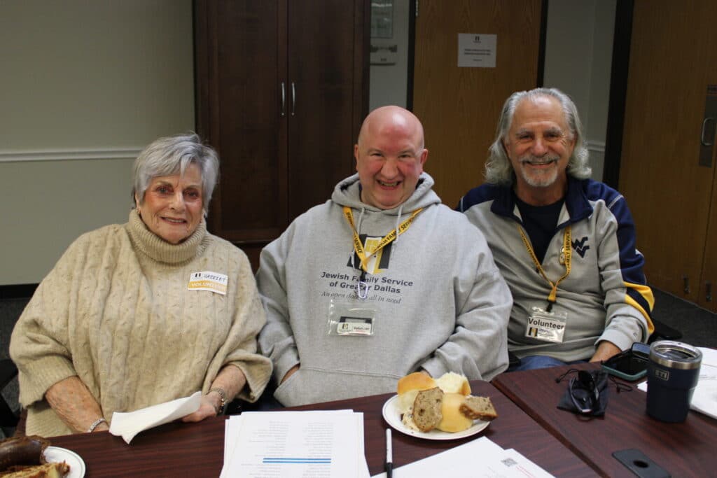 Three smiling older adults sit at a table with bread and papers, wearing volunteer name tags and lanyards.