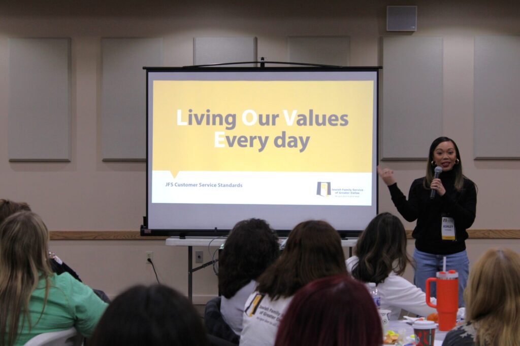A woman speaks to an audience in front of a screen displaying "Living Our Values Every day.