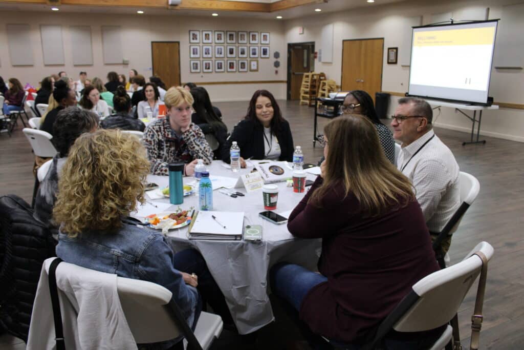 A group of people sit around a table talking at a workshop in a conference room with a presentation screen in the background.