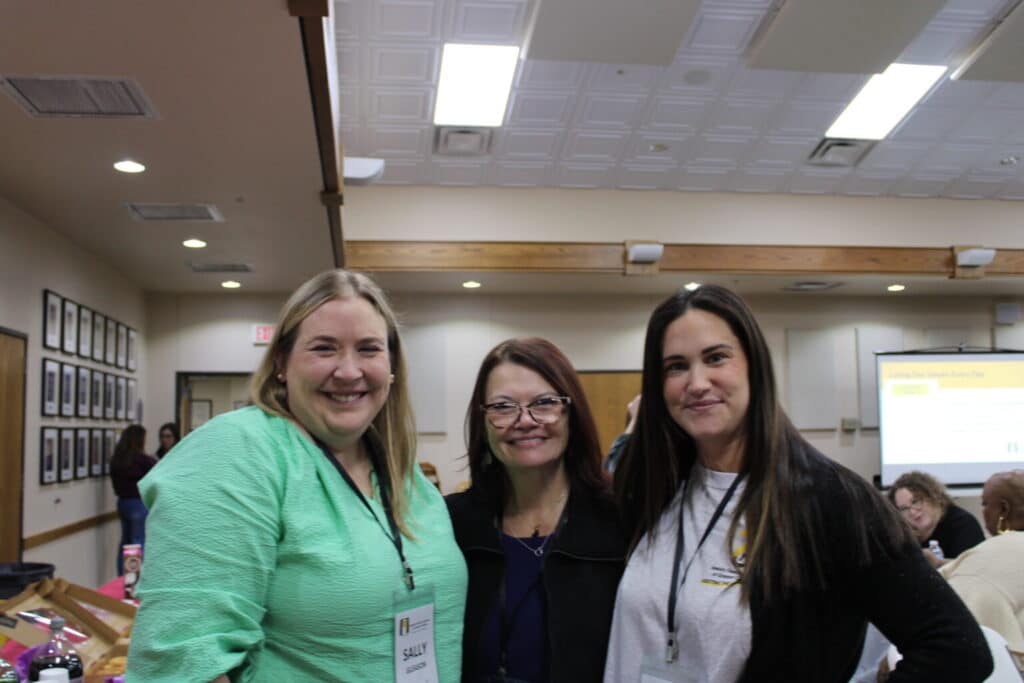Three women smiling and posing together in a conference room, with people and a presentation screen in the background.