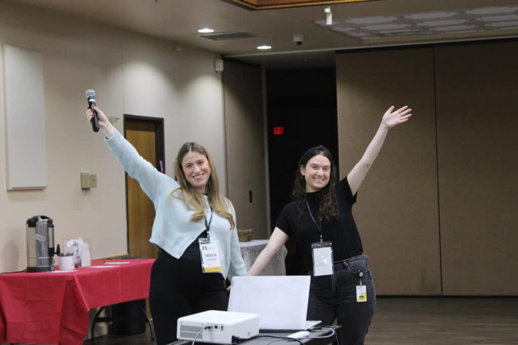 Two smiling women stand by a laptop, one holding a microphone, both with arms raised in a welcoming gesture.