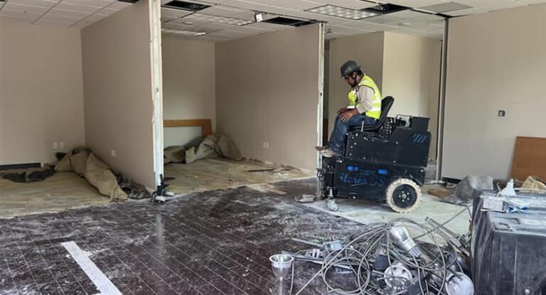 A construction worker operates machinery in a partially demolished office space with exposed floors and walls.