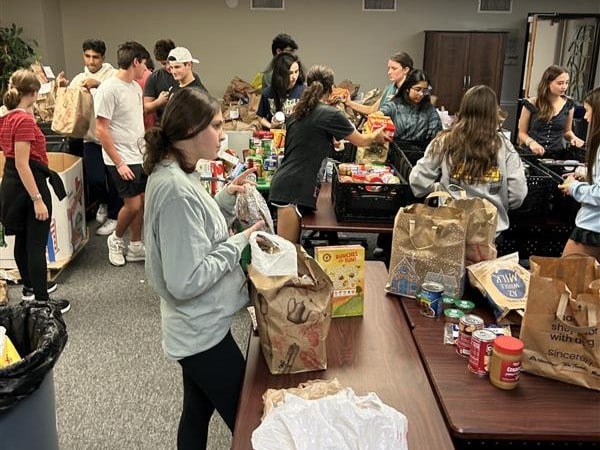 A group of people sorts and packs canned food and groceries into bags on tables in a busy room.